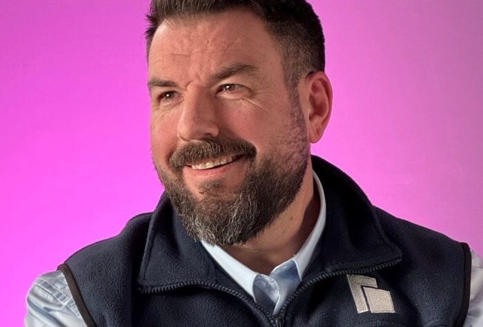 Lee Witts in a pale blue shirt and Brookbanks gillet sat in front of a pink bacground