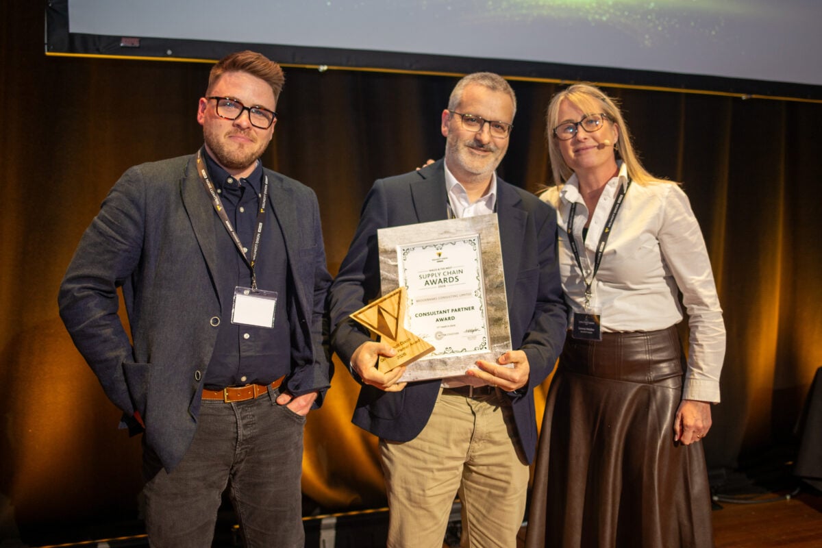 Tom Quaife-Jones and Chris Vivian on stage at the Willmott Dixon Supplier Chain Awards 2026 representing Brookbanks. Chris is stood in the middle and is holding a certificate and small wooden trophy.