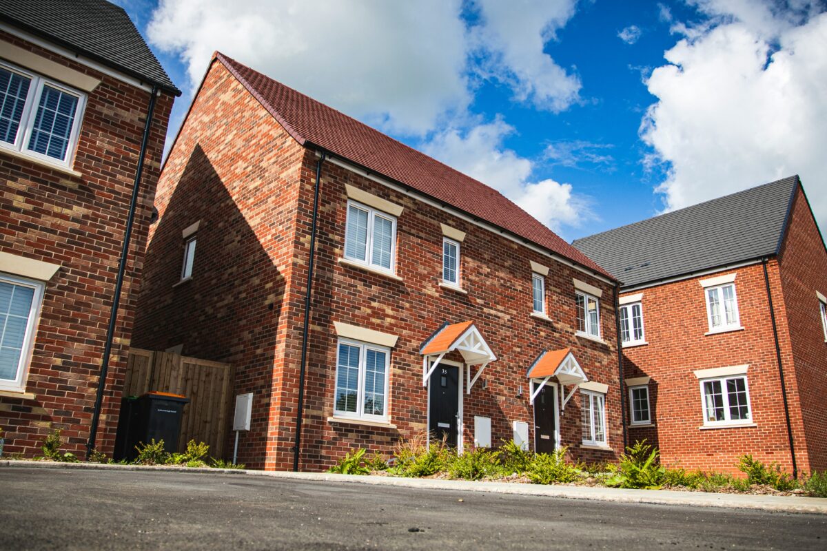 A red brick semi detached house in a residential area. It has a grey driveway with greenery in the front garden