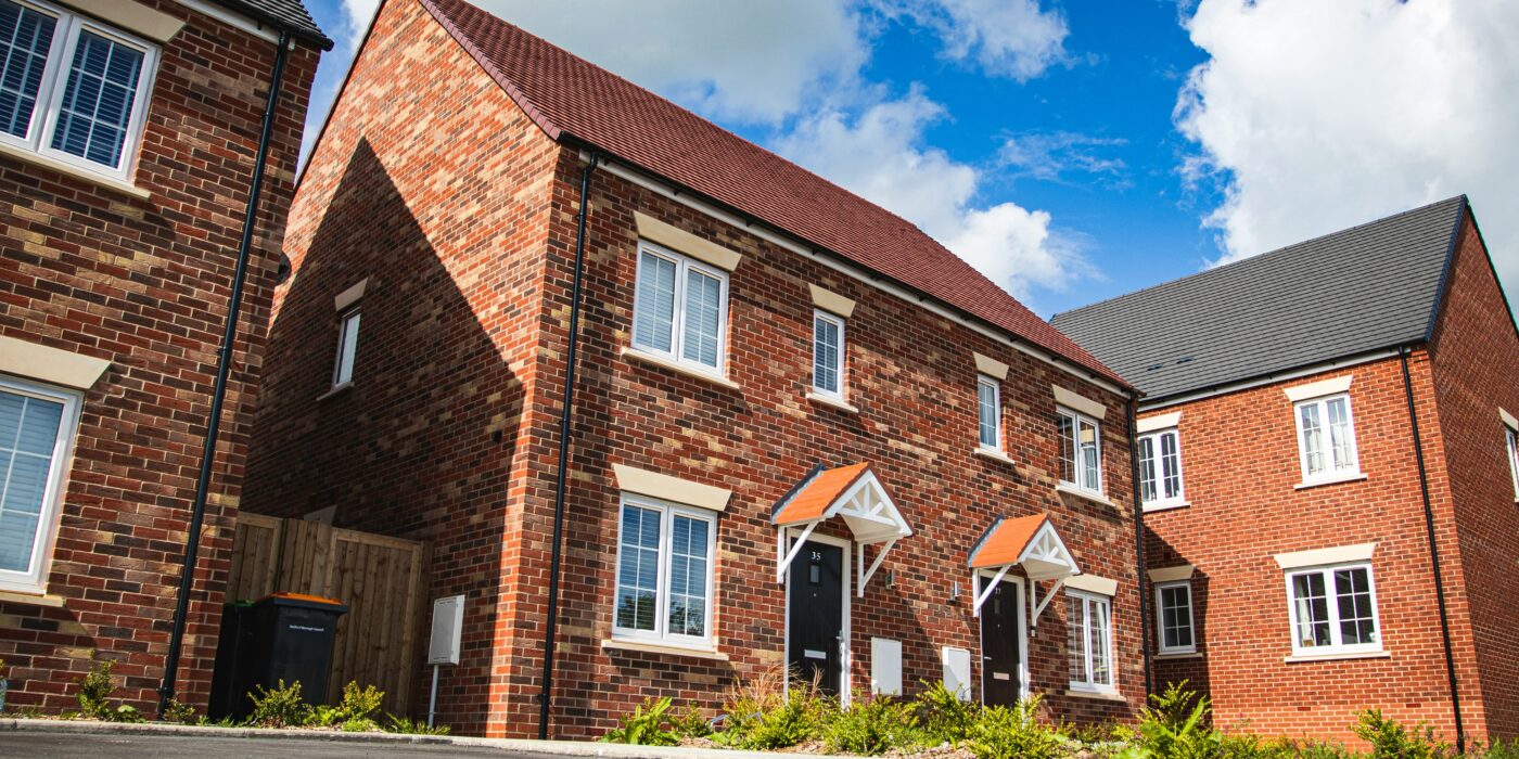 A red brick semi detached house in a residential area. It has a grey driveway with greenery in the front garden
