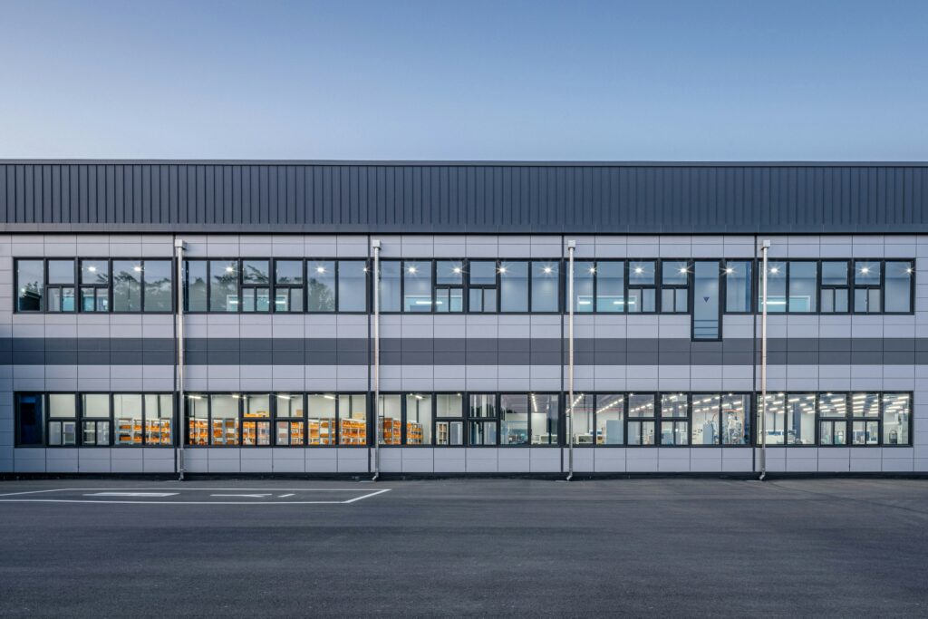 Front elevation of a modern industrial and office building with large windows, illuminated interiors, and an empty parking area in the foreground.