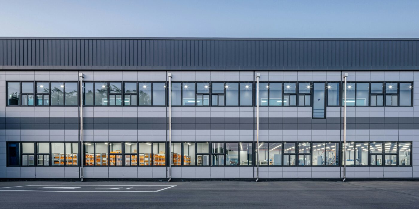 Front elevation of a modern industrial and office building with large windows, illuminated interiors, and an empty parking area in the foreground.