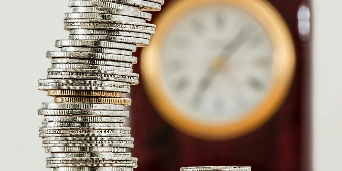 A close-up image of stacked coins with a blurred clock, symbolizing time and money relationship.