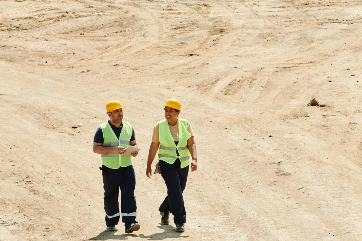 Two workers in safety vests and hard hats walking on a sandy construction site.