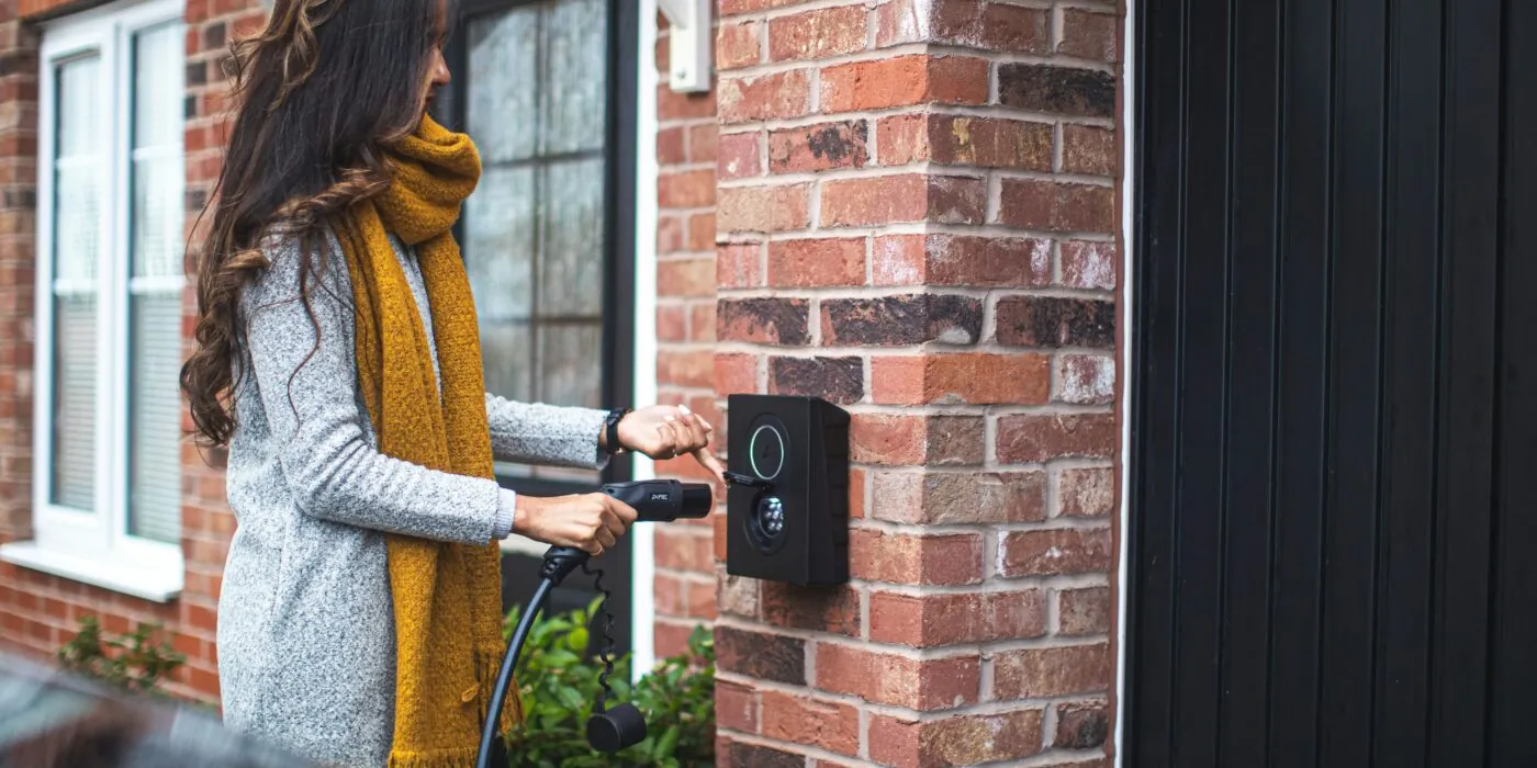 A woman stood outside her house taking off an electric vehicle charger off its hook. It is attached to a brick wall on the outside of the building.